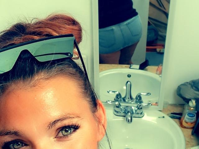 SexyGreenEyes69, avec ses fesses visibles dans un short bleu, se reflète dans le miroir, portant des lunettes de soleil sur la tête, devant un lavabo de salle de bain et des articles de toilette, montrant ses yeux verts et son chignon brun..