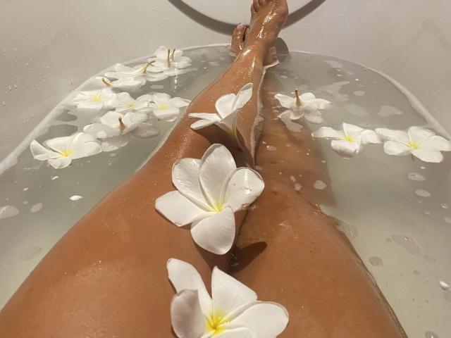 ReineDuVice's legs in a bathtub with white plumeria flowers floating on the water. Her tan skin is wet, and the flowers are arranged along her thighs.