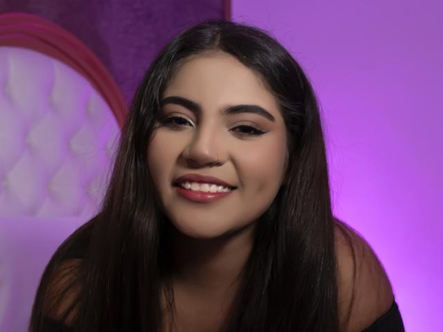 Portrait of VictoriaRosel with long, straight brown hair, fair skin, and dark eyebrows. She has a small nose and full lips, smiling slightly. She wears a black off-shoulder top. The background has a purple gradient with a white, tufted headboard on the left and a pink curved arch. Her makeup includes subtle eyeliner and pink lipstick. The image has a soft, even light, highlighting her facial features. The composition centers VictoriaRosel's face, with a slight upward angle. The overall style is clean and modern.