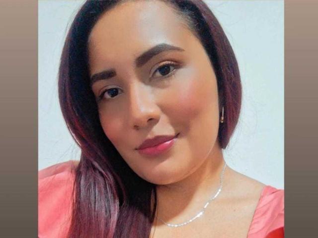 AlizWolf with medium brown skin, dark red hair, pink lipstick, subtle makeup, and a small smile, wearing a red top and silver necklace, looking at the camera.