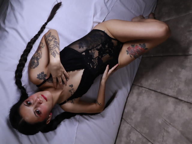 Yliria with long braids, black lace bodysuit, tattoos on arms and thigh, lying on white bed, legs spread, looking up, hand on chest, showing her tits and pussy.