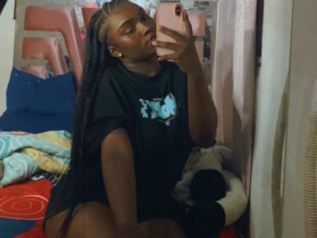 LoveeBonyyJuices mit dunkler Haut, langen Zöpfen, einem schwarzen T-Shirt und einem rosa Handy, macht ein Selfie vor einem Spiegel und zeigt ihren Hintern, während sie auf einem Bett mit bunten Kissen sitzt..