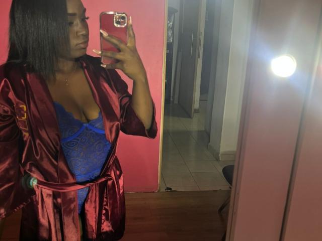LoveeBonyy, vêtue d'une robe en satin rouge, dévoilant de la lingerie en dentelle bleue avec un décolleté plongeant, tenant un téléphone rouge, autoportrait devant un miroir, cheveux noirs courts, lumière réfléchie sur le sol..