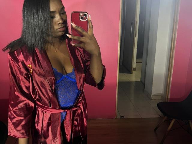 LoveeBonyyJuices with dark skin, long black hair, wearing a red satin robe, blue lace lingerie, holding a red phone, showing her tits in a mirror selfie.