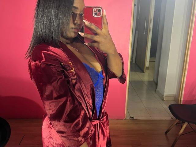 LoveeBonyyJuices, avec de longs cheveux noirs, portant une robe en satin rouge et un soutien-gorge en dentelle bleu, se prend en photo devant un miroir avec un téléphone rouge, montrant ses seins, dans une pièce aux murs roses..