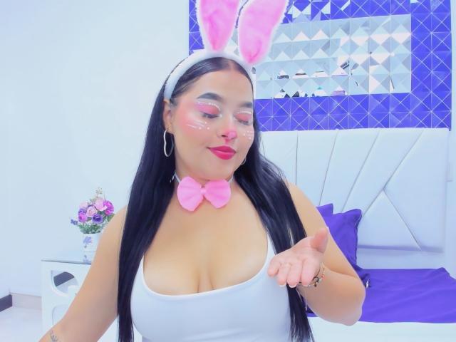 CandasLeroy con orejas de conejo rosas, diadema blanca, lazo rosa y camiseta blanca, señalando a la cámara, mostrando senos grandes, cabello negro largo, sombra de ojos rosa y lápiz labial rojo..