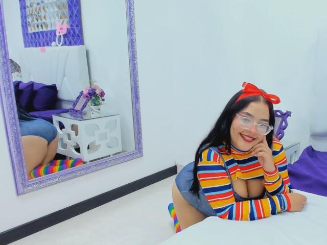 CandasLeroy with long black hair, red bow, glasses, and striped top, showing big boobs, in denim shorts, rainbow knee socks, smiling, leaning on bed, reflected in mirror.