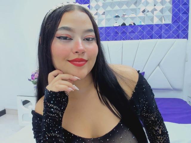 CandasLeroy has black hair, red lipstick, and wears a sparkly black top, showing off her tits. She's smiling with a hand on her chin, in a bedroom.