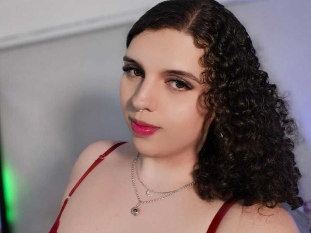 CatleyaNoire, con cabello castaño rizado, piel clara, lápiz labial rojo, vistiendo un sujetador rojo y un collar de plata, mirando a la cámara con una ligera sonrisa..