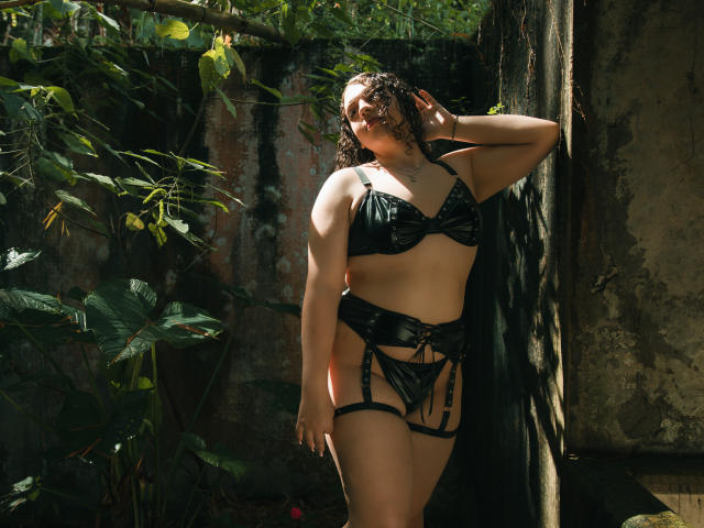Una Curvy CatleyaNoire con capelli scuri e ricci è in piedi, indossando lingerie in pelle nera, bretelle e lacci. La luce del sole filtra attraverso le foglie verdi, e una mano le tocca la testa, mostrando il suo seno prosperoso e i glutei..
