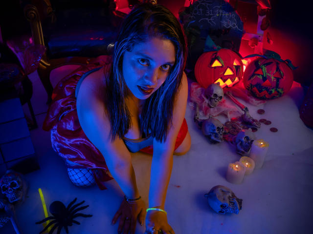 ValeriaDeleon, com cabelos loiros molhados, ajoelhada, vestindo um vestido vermelho e meias de rede, cercada por velas brilhantes, caveiras e abóboras de Halloween, olhando para cima com iluminação intensa em azul e vermelho..
