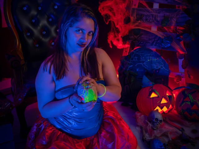 ValeriaDeleon, con la piel iluminada en azul, sosteniendo un recipiente verde brillante, vestida con un top gris sin tirantes y una falda roja, sentada en una habitación oscura y espeluznante con una calabaza encendida..