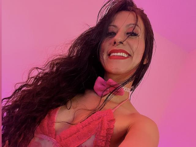 AmaraMirage with long, wet, dark brown hair, smiling, wearing a pink lace halter top and matching pink bowtie, pink lighting, showing her tits and red lipstick.