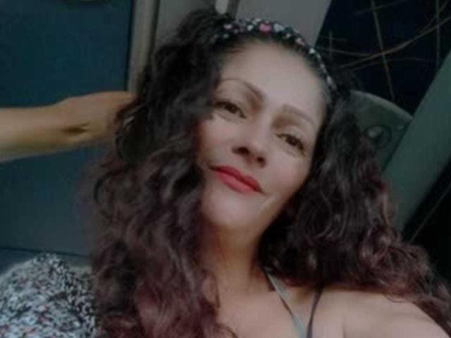 A MegaanTaylor69 tem cabelos escuros e cacheados, uma tiara com flores, batom vermelho e está sorrindo, com a mão no cabelo, mostrando seus ombros nus e uma blusa estampada..