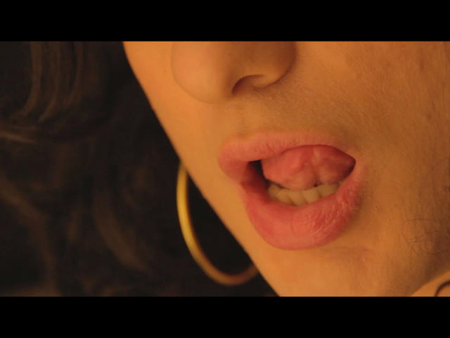 Close-up of ChantalCross-hot's face, focusing on her slightly parted pink lips with a visible tongue. She has a small, upturned nose and a subtle, natural skin tone. A gold hoop earring is on the right side of her face. Her dark, wavy hair is partially visible on the left. The image uses warm, soft lighting, highlighting her lip texture and earring. The overall composition is intimate and detailed.