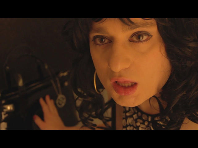 Close-up of ChantalCross-hot with wavy black hair and pale skin, holding a black camera with a circular logo. She has dark, arched eyebrows, light brown eyes, and slightly parted red lips. She wears large gold hoop earrings and a black lace top. The camera is in her right hand, positioned near her face. Warm, yellowish lighting creates a soft, intimate atmosphere. The focus is on her face and the camera.