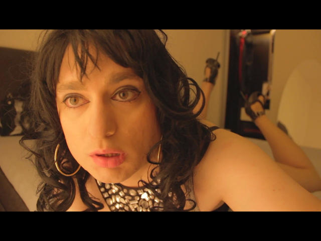 Close-up photo of ChantalCross-hot with wavy black hair, green eyes, and pink lips. She wears a silver sequined necklace and large hoop earrings. She's lying on a bed with legs bent at the knees, wearing black high heels. The warm lighting highlights her pale skin and slight redness on her cheeks. Her gaze is direct, and she looks slightly to the side. The image has a soft focus with a slight depth of field.