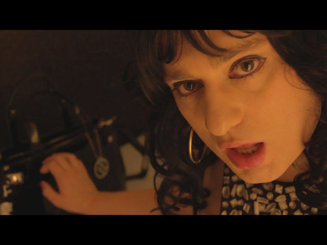 Close-up of ChantalCross-hot with curly black hair and bangs, wearing a black-and-white patterned top, and large gold hoop earrings. Her brown eyes are wide and slightly squinted, with dark eyeliner accentuating her gaze. Her pink lips are slightly parted. She holds a black camera with a round lens in her right hand. The warm, low-light setting creates a shadowy effect, highlighting her facial features. The image has a cinematic, slightly gritty style. Her expression is intense and focused.