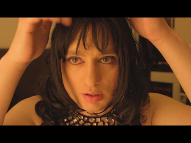ChantalCross-hot has wavy black hair, pale skin, and green eyes. She's wearing a metallic chain choker. Her hands are in her hair, and her mouth is slightly open with pink lips. The image is close-up, focusing on her face. Warm, yellowish lighting enhances her skin tone. The background is blurred. The composition centers on her facial expression and hair. The image has a letterbox format with black bars at the top and bottom.