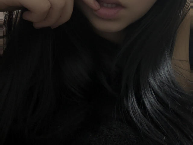 Close-up of Kkatalina's face and upper chest. She has long, straight, black hair that partially covers her face. Her lips are slightly parted, showing a small portion of her upper teeth. Her right hand is near her face, with her index finger gently touching her lower lip. The lighting is low, creating a shadowy effect. The image focuses on her hair and lips, with a subtle texture in the dark hair. Her skin is light. The composition is intimate and somewhat mysterious.