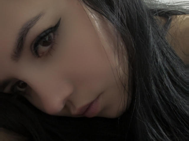 Kkatalina's face, showing dark eyebrows, black eyeliner, and long black hair, with a pouty expression, is the focus of this tight shot. Her brown eyes look slightly downward.