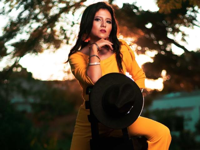 RenataRivas with dark hair, wearing a yellow dress, black hat, and bracelets, holding her chin, standing in front of a blurred sunset, with trees in the background.