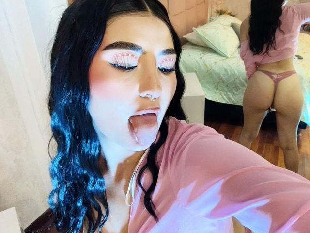 MaddisonCarter with black hair, pink makeup, tongue out, wearing pink top and pink thong, showing her ass in mirror reflection. Her eyes are closed. She's on a bed.