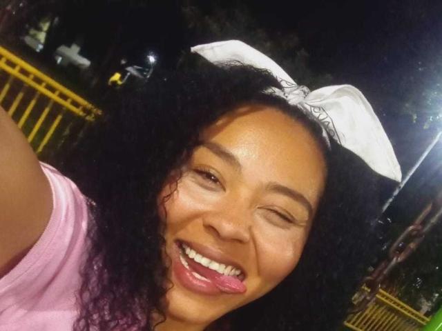 GenesisReyes with dark curly hair, winking, sticking out tongue, wearing white nurse cap, pink shirt, and smiling, night-time selfie.