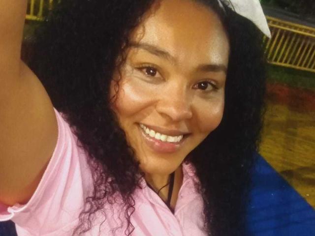 GenesisReyes with curly black hair, smiling with white teeth, wearing a pink shirt, raising her arm for a selfie, brown eyes, and a light brown skin tone.