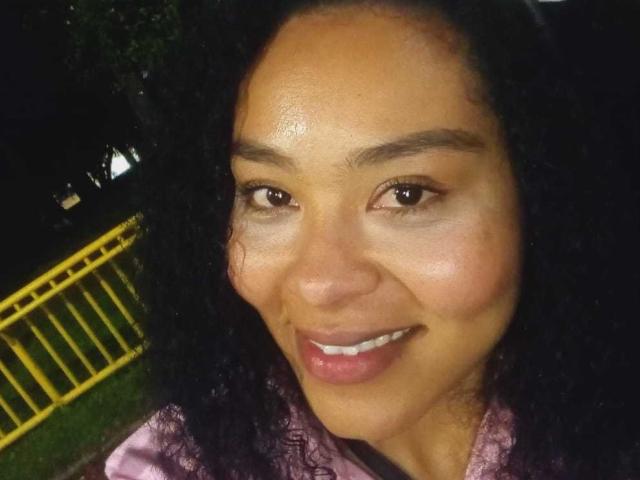 GenesisReyes has dark curly hair, brown eyes, and a small smile. She's wearing a pinkish shirt, with a shiny, slightly sweaty face. Her tits are not visible.