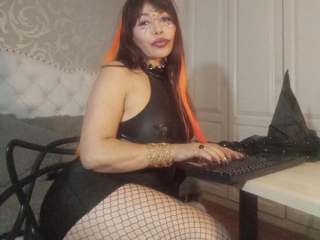 LilithCalderon with orange hair, black leather dress, fishnet stockings, gold bracelet, and makeup, sitting at a laptop, showing her ass, on a tufted chair, in a room.