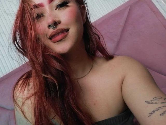 Samara, une jeune femme aux cheveux roux, avec un anneau au nez et un tatouage sur son bras droit, la langue tirée, montrant ses épaules nues et un haut gris sans bretelles. Elle sourit à l'appareil photo..