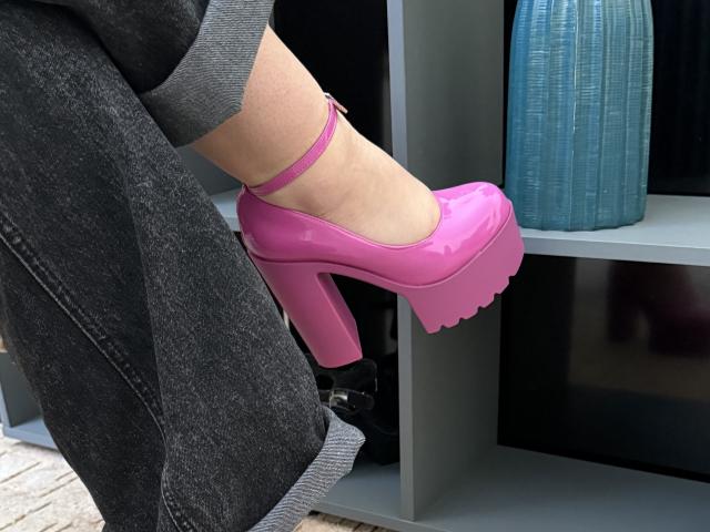 Le scarpe rosa, lucide e con zeppa alta di SintiaMaers, con suole spesse e ondulate, sono appoggiate su uno scaffale grigio, accanto alle sue gambe nere, con risvolto e rivestite di denim..