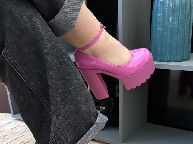 Les élégantes chaussures à talons roses de SintiaMaers, avec une semelle épaisse et une bride à la cheville, sont portées par une jambe dont la chaussette est grise, et qui porte un jean foncé. Son pied est levé, ce qui permet de voir le talon..