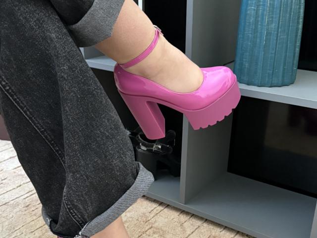 Close-up of a foot in a bright pink, high-platform, stiletto heel. The heel is chunky with a scalloped edge, and a small ankle strap is visible. SintiaMaers is wearing dark blue, cuffed jeans with the bottom of the jeans rolled up. The foot is slightly raised, showing the sole of the heel. A blue vase is on a gray shelf in the background. The texture of the jeans is rough, contrasting with the shiny, smooth surface of the heel. The image focuses on the vibrant color and bold design of the shoe.