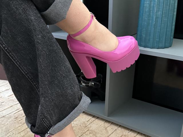 SintiaMaers' rosa, glänzende, plattformhohe Schuhe mit einer dicken, schuppigen Sohle sind angehoben und zeigen ihren Knöchel sowie ihre dunklen Jeans mit Bund, vor einem grauen Regal mit einer blauen Vase..