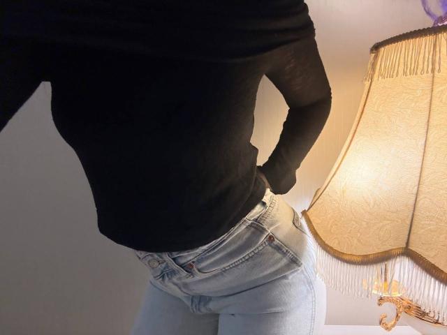 KaiaLein's black top and light blue jeans show a slightly curved ass and hands on her hips, with a lamp in the background.