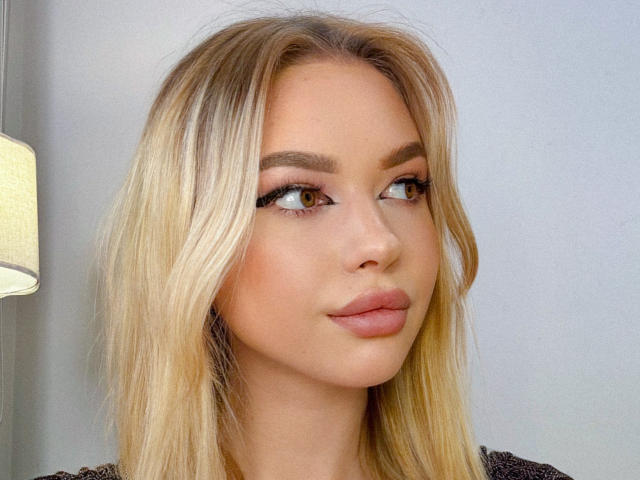 Close-up portrait of Adellume with light blonde hair, slightly wavy, and parted to the side. She has thick, dark eyebrows, long, curled eyelashes, and brown eyes looking to the right. Her lips are full, with a subtle pinkish-brown hue. She's wearing a dark, textured top. A soft, yellow lamp is partially visible on the left. The skin tone is fair with a slight natural glow. The composition is centered on Adellume's face, emphasizing her facial features. The image has a clean, modern look with a focus on natural beauty.