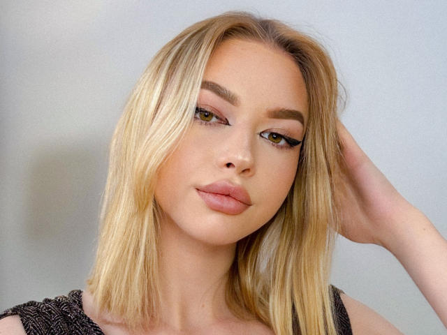 Adellume hat blondes, glattes Haar, braue Augenbrauen und trägt Eyeliner mit langen Wimpern. Sie schaut mit einem leichten Lächeln in die Kamera, eine Hand in ihrem Haar..