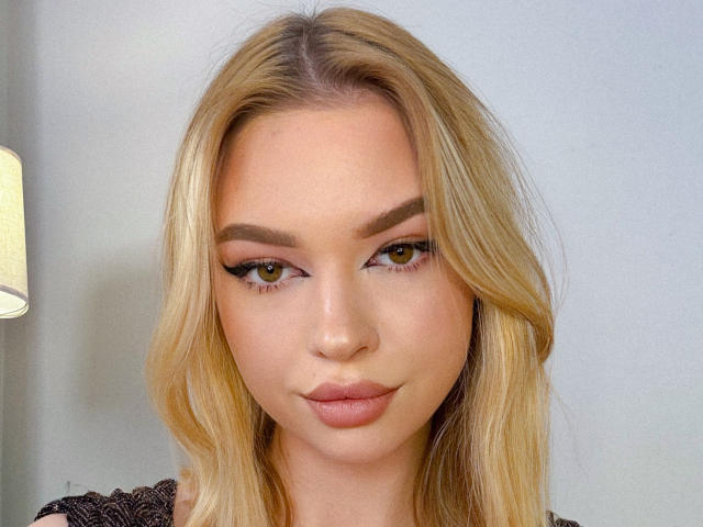 Close-up portrait of Adellume. She has blonde, wavy hair that falls past her shoulders. Her eyebrows are thick and dark, contrasting with her fair skin. She has yellow-green eyes with pronounced eyeliner and long lashes. Her lips are slightly parted, showing a subtle, natural pink color. She's wearing a black, lace-patterned top. A small portion of a beige lamp is visible on the left. The background is a plain, light gray wall. The image has a smooth, realistic texture.