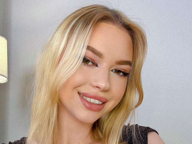 Close-up of Adellume with blonde hair, fair skin, and brown eyes. She has a subtle smile with pink lipstick. Her eyebrows are neatly shaped, and she has long, slightly wavy hair. She's wearing a dark, textured top. The lighting is soft, highlighting her facial features. The background is a plain, light gray wall. The image is sharp with a realistic style. Adellume's makeup is light, with a focus on her eyes and lips.
