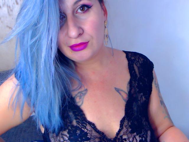 LilithGreenn with blue hair, pink lipstick, black lace top, tattoos on chest, and gold earrings, showing off her tits. She's smiling and looking at the camera.