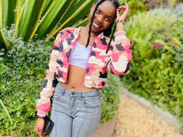Niiia stands in a garden, smiling at the camera. She wears a pink, black, and beige camouflage jacket, slightly open to reveal a white crop top. Her blue jeans are high-waisted, and she holds a black clutch in her right hand. Her braids are neatly styled, and she has a bracelet on her left wrist. The greenery behind her includes large leaves and various plants. Niiia's skin is dark, and she has a slim build. The image is brightly colored and focused on her upper body.