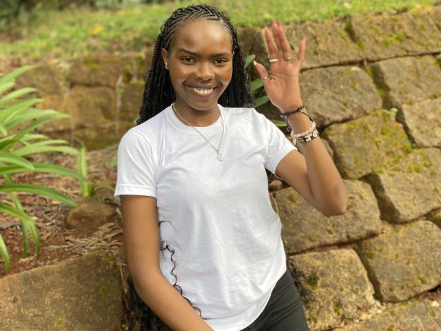 Niiia with dark skin, braided hair, white t-shirt, black pants, smiling, waving, rock wall, green plants, bracelet, necklace, standing outside, looking happy.