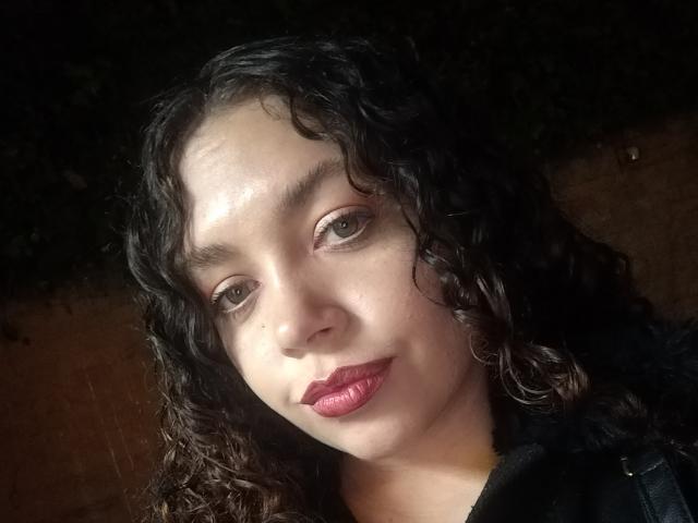 Close-up of RubbyStrongX with curly black hair, light brown skin, and dark brown eyes. RubbyStrongX has glossy, pink lipstick and a slight smile. The light source is from the upper left, highlighting her face and creating shadows on the right. She's wearing a black garment, and the background is dark with a hint of green foliage. The texture of her hair is wavy and slightly shiny. The overall composition is centered and slightly tilted.