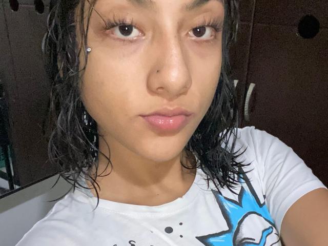 SheaCum has wet black hair, a nose piercing, and glossy pink lips. She's wearing a white shirt with a blue and black graphic, showing a neutral expression.
