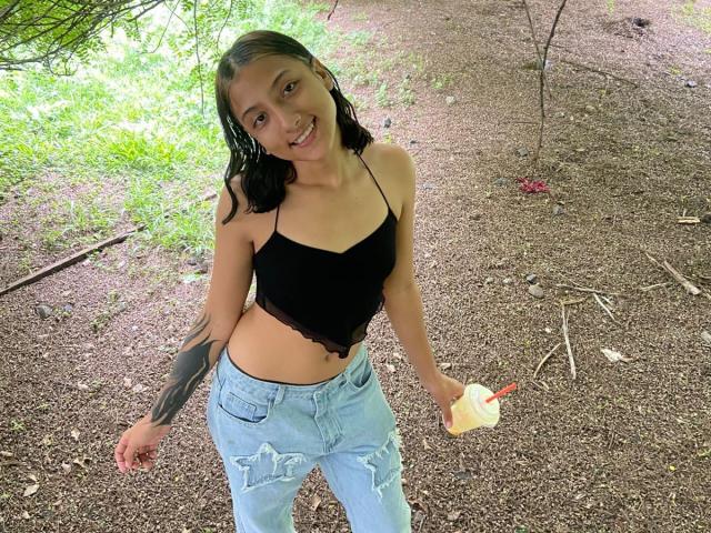 SheaCum with wet black top, ripped blue jeans, holding ice cream, tattoo on right arm, smiling, brown hair, small tits, standing on dirt path, holding yellow ice cream.
