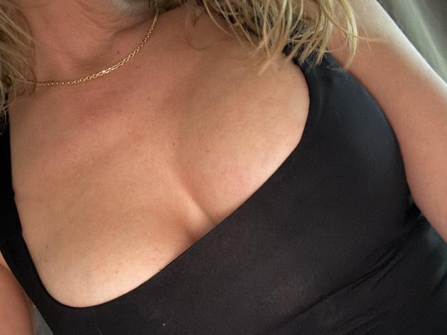HotFrenchy69's tits in a black top, showing a bit of cleavage, with a gold chain necklace and blonde wavy hair. Her skin is light with some freckles.