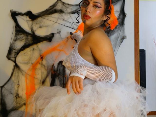 AmaraMartelli sits in a semi-profile position, wearing a white lace dress with a fluffy tulle skirt and fishnet arm sleeves. Her curly hair is adorned with an orange ribbon. She has red lipstick and pink eyeshadow, with a subtle blush. The background features abstract black and orange splashes on a white wall. Her right arm rests on her knee, and she gazes slightly to the side. The overall composition is bold and striking.