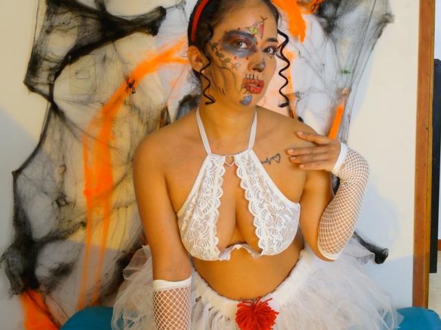 AmaraMartellI, a woman with dark curly hair, wears a white lace bralette exposing her medium-sized breasts, and white fishnet arm sleeves. She has face paint with colorful patterns, including red lips and blue eyes. She sits against a black and orange abstract backdrop. She wears a white tulle skirt with a red flower. Her right hand is raised, touching her chest. The image is vivid, with a focus on her detailed face paint and lace attire.